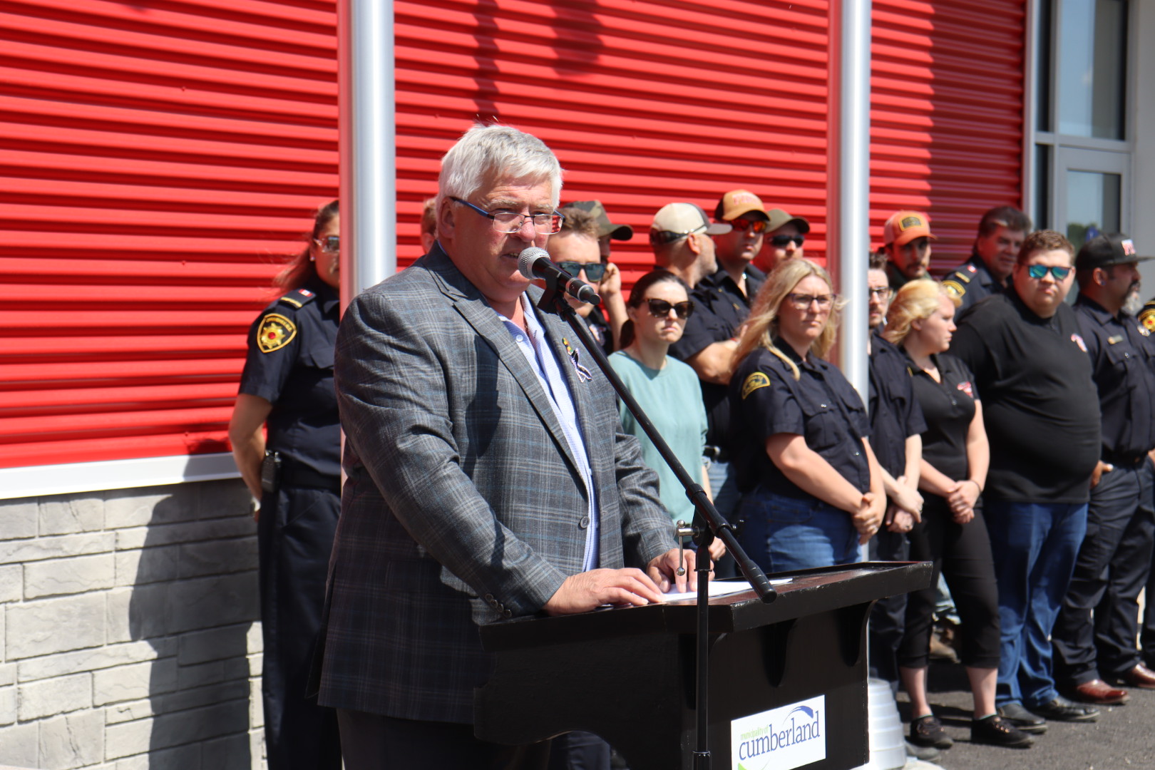 A man stands at a podium speaking while a group of people stand in behind.